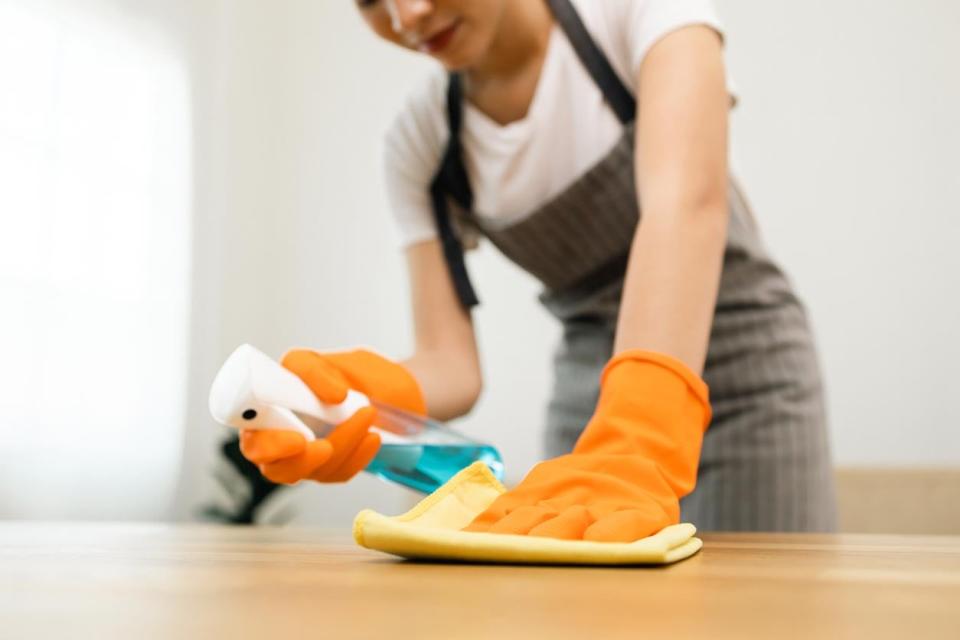  A person in orange gloves sprays a cleaning solution on a table and wipes it down with a yellow cloth.