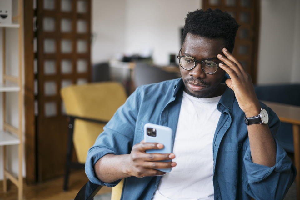 Man in glasses and a casual outfit looks at his phone with a thoughtful expression, sitting in an office space