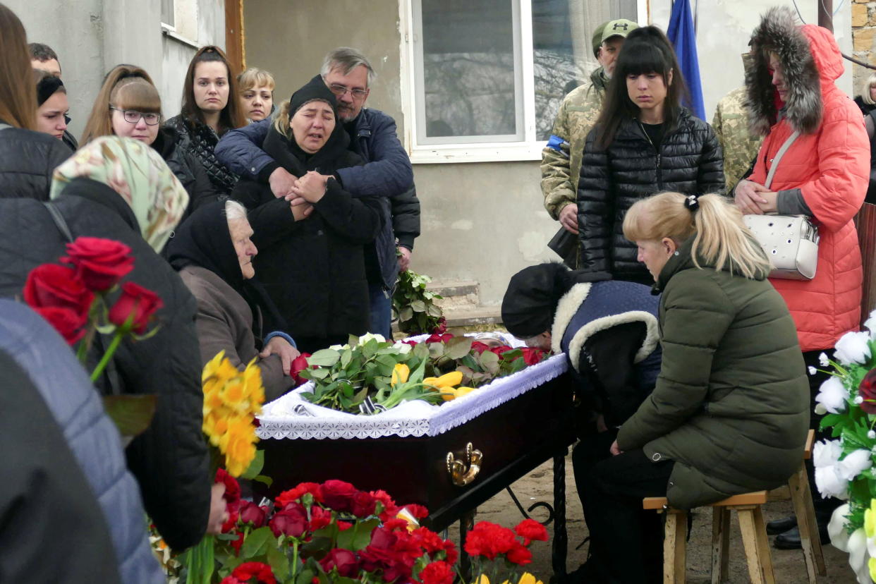 A middle-aged woman, embraced from behind by a man with graying hair, by cries over a coffin covered with flowers, as other mourners look on.