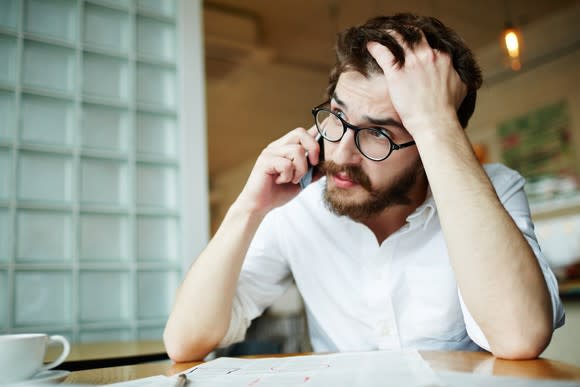 Young male adult on phone sitting at a table with spread-out papers and running his hand through his hair.