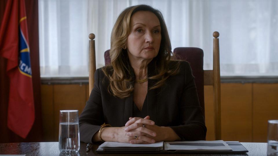 Close up of an adult woman sitting at a desk with her hands clasped in front of her. She looks powerful with her glaring expression and smart blazer.