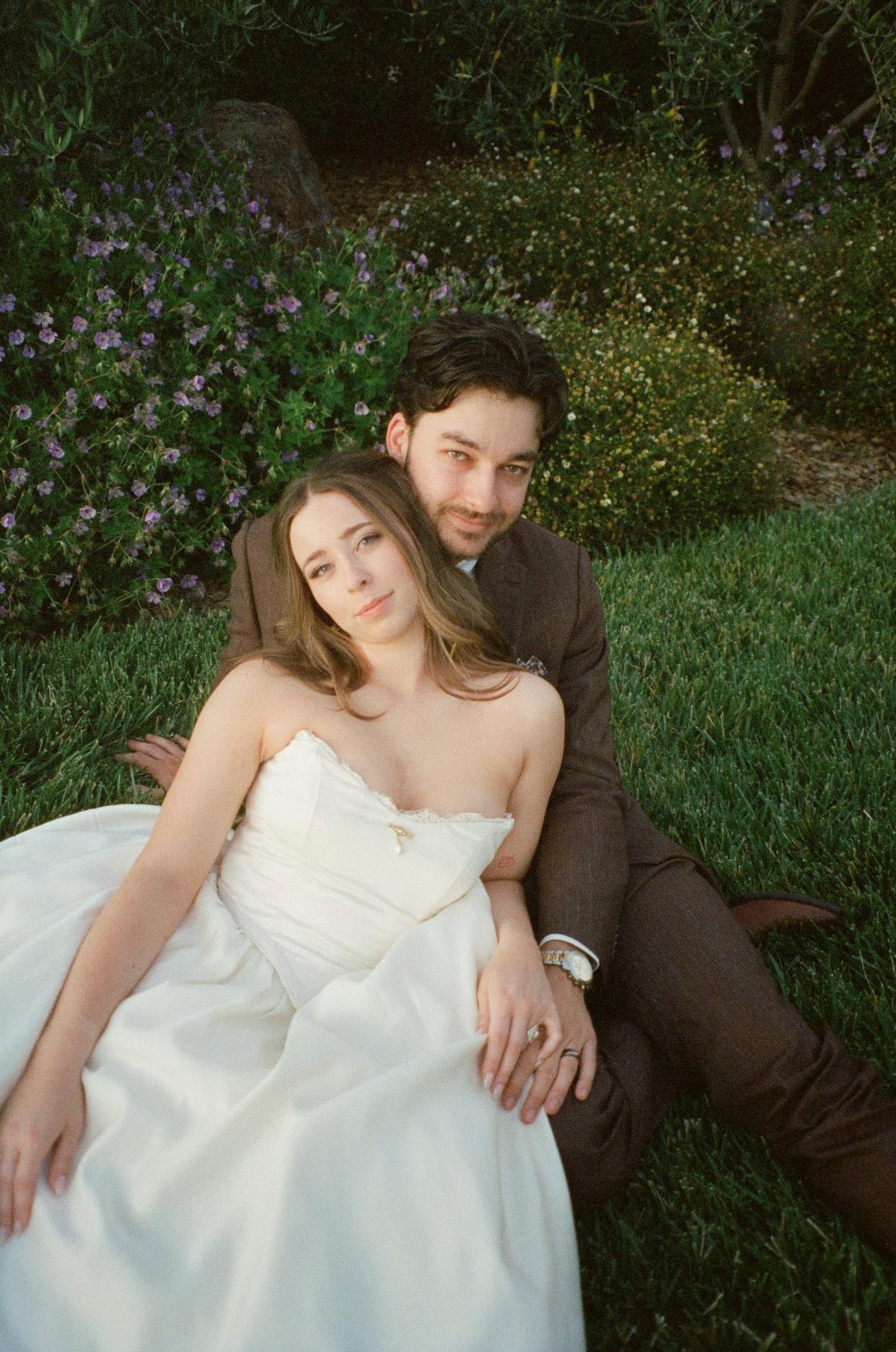 A bride sits in front of her groom and leans back on him in a grassy field.