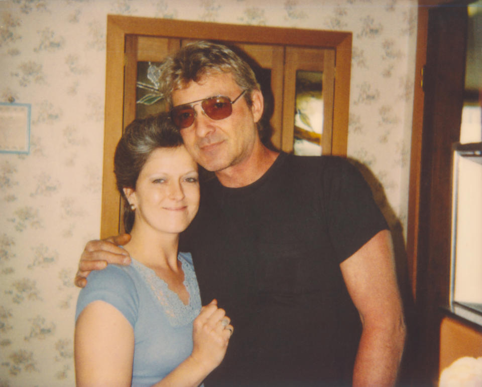 Two people smiling and embracing for a photo in a room with floral wallpaper