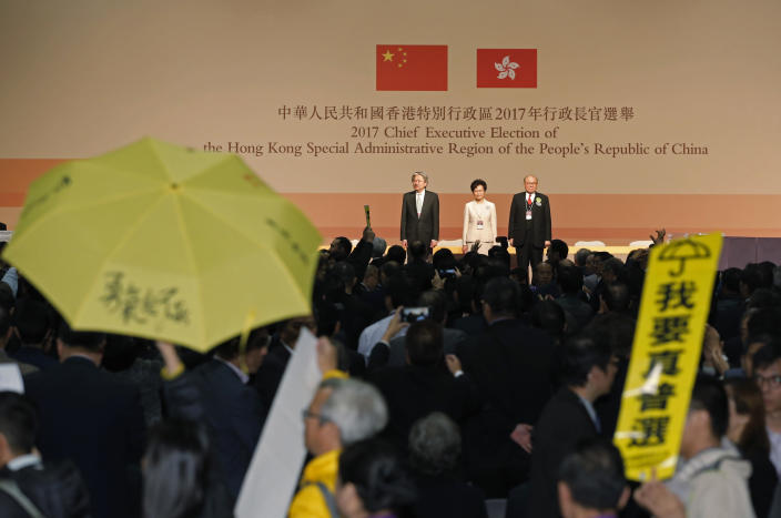 FILE - A protester raises an umbrella and placard reading: "I want genuine universal suffrage" to protest as former Hong Kong Chief Secretary Carrie Lam, center, declares her victory in the chief executive election of Hong Kong while former Financial Secretary John Tsang, left, and retired judge Woo Kwok-hing stand with her in Hong Kong on March 26, 2017. (AP Photo/Kin Cheung, File)
