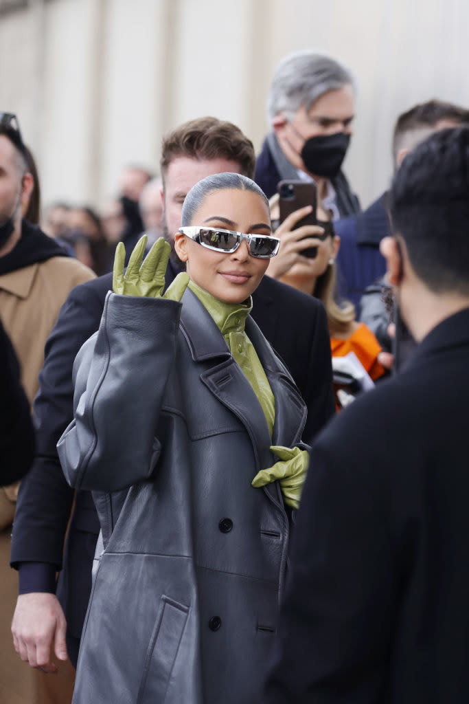 Kim, wearing sunglasses, a leather trench coat, and gloves, waves outside the Prada show during the 2022 Milan Fashion Week