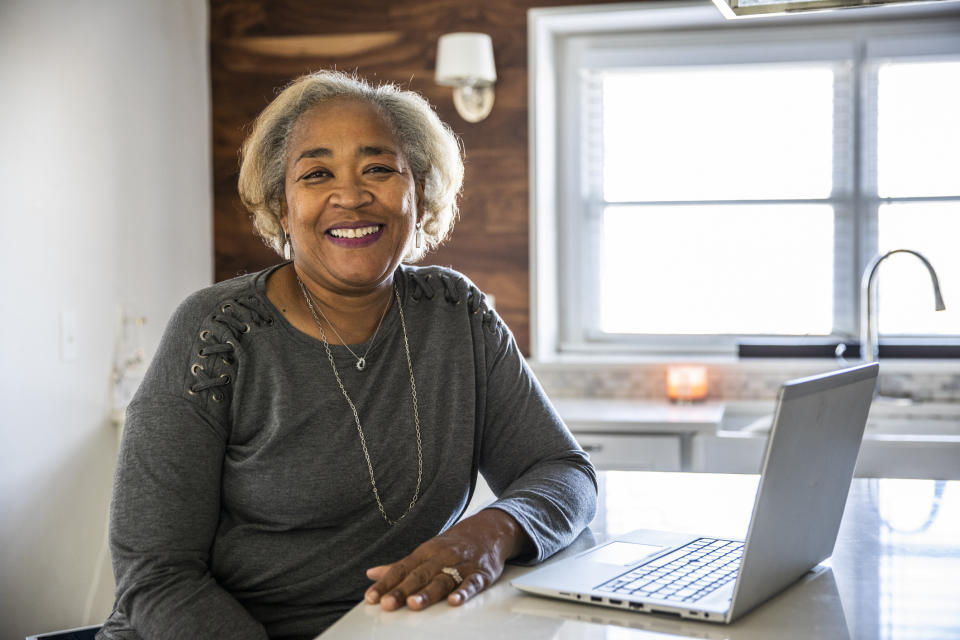 Woman smiles at camera with a laptop on a table in a home setting. She wears a casual top with shoulder details