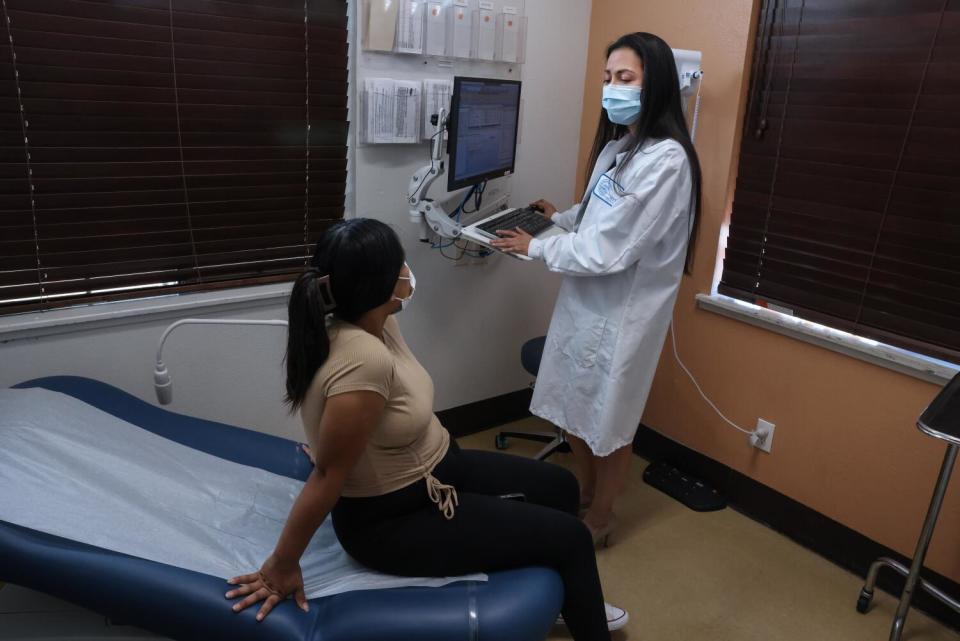 A masked woman with long dark hair, wearing a white coat, right, speaks to a woman sitting in an examination chair