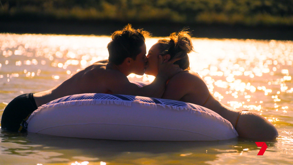 Will shares a kiss with a woman in a lake over an inflatable ring, sunset drenched water