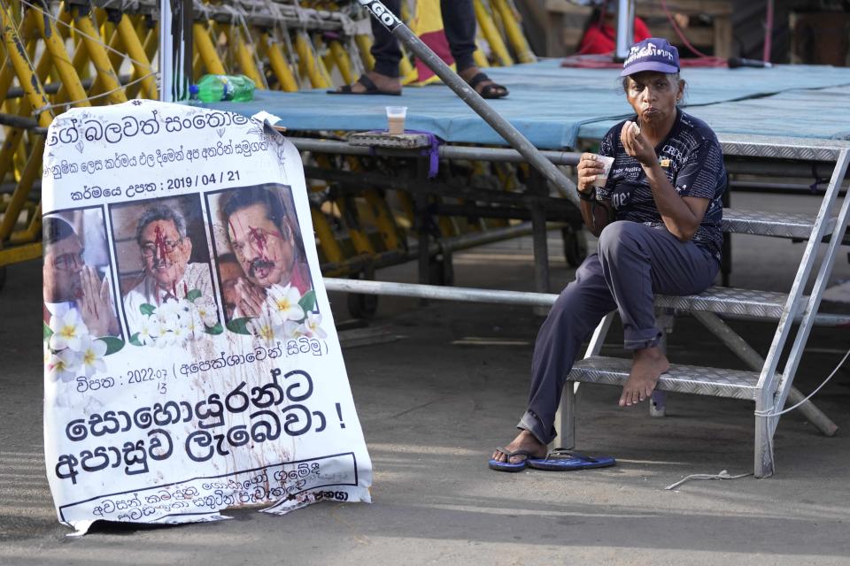 A protester drinks a cup of tea as she sits by a defaced poster carrying portraits of ousted president Gotabaya Rajapaksa, center, and his brothers at the entrance to president's office in Colombo, Sri Lanka, Friday, July 15, 2022. Protesters retreated from government buildings Thursday in Sri Lanka, restoring a tenuous calm to the economically crippled country, and the embattled president at last emailed the resignation that demonstrators have sought for months.(AP Photo/Eranga Jayawardena)