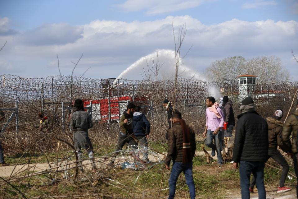 Greek firefighters spray water as migrants stand near a border fence on the Turkish side during clashes with the Greek riot police at the Turkish-Greek border in Pazarkule, Edirne region, on Saturday, March 7, 2020. Thousands of refugees and other migrants have been trying to get into EU member Greece in the past week after Turkey declared that its previously guarded borders with Europe were open. (AP Photo/Darko Bandic)