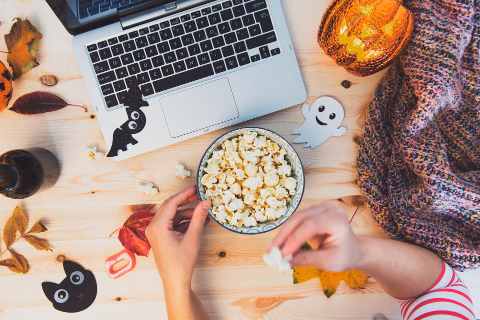 Top view Woman eating popcorn while watching Halloween movie. Laptop, popcorn bowl, pumpkin, bats, ghost silhouettes, sweets, fall leaves, warm plaid on wooden background. Cozy and safe holiday party