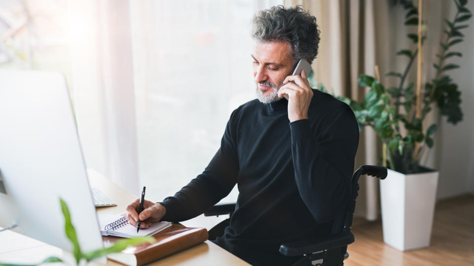 Handsome mature man in wheelchair with smartphone in home office, making a phone call.