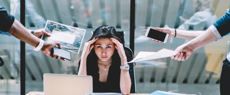 Woman working at desk, looking stressed while people hand her phones and paperwork