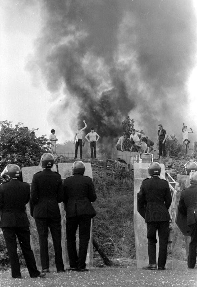 Police watch as pickets face them against a background of burning cars at the Orgreave coke works in June 1984