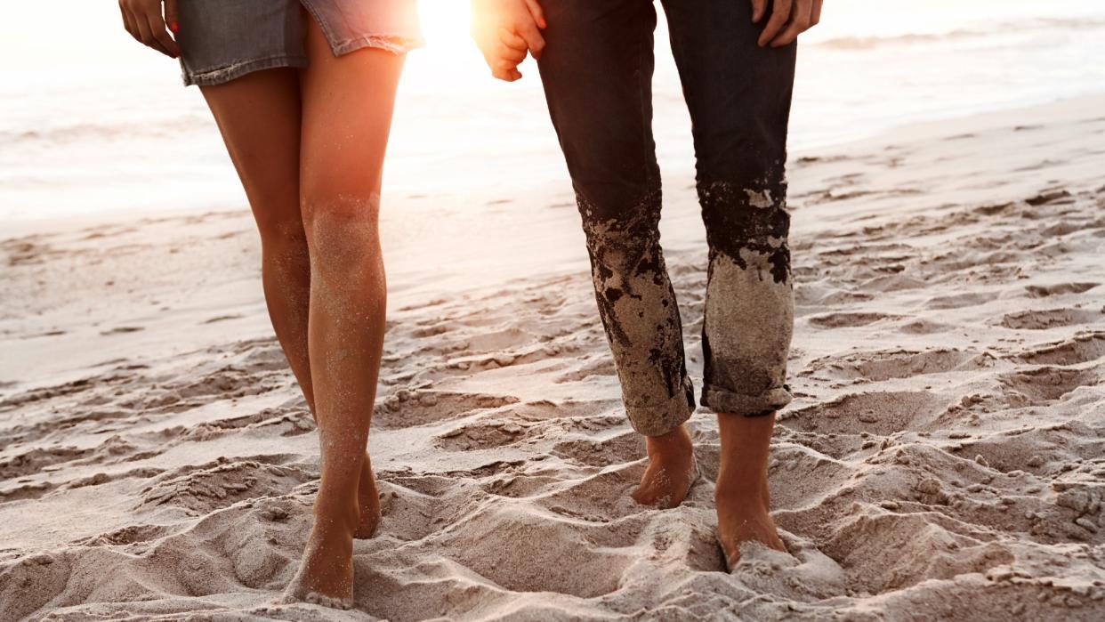  Couple holding hands and walking barefoot on the beach as the sun sets in the background. 