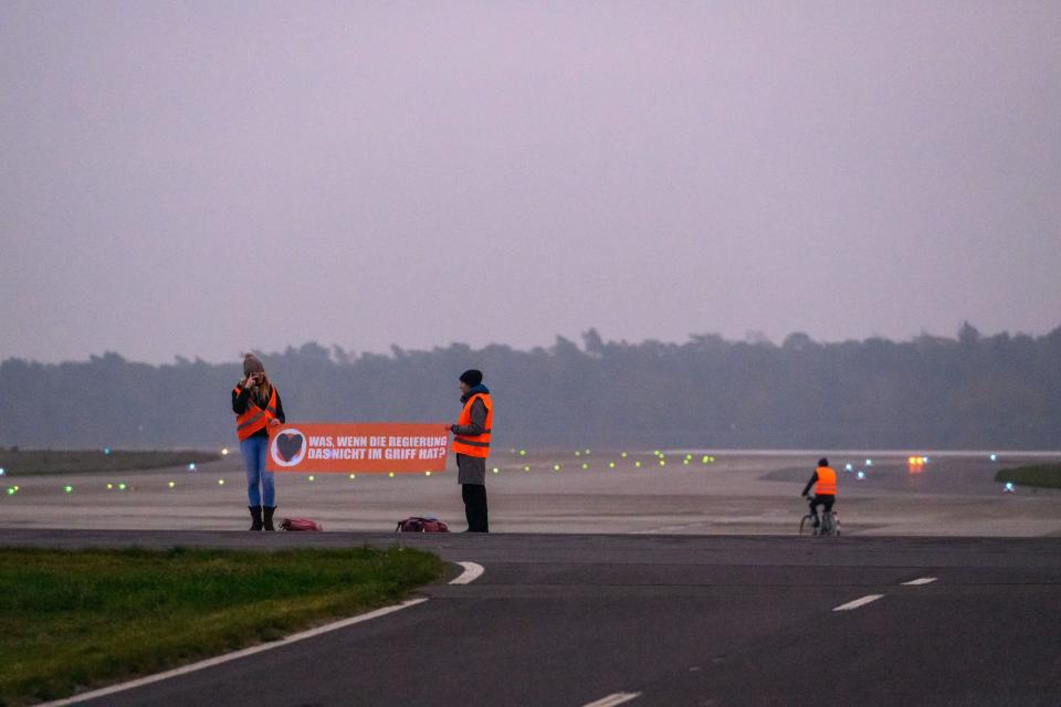 Aktivisten vom Aufstand der Letzten Generation auf der Landebahn des BER mit Fahrrad und Transparent mit der Aufschrift "Was, wenn die Regierung das nicht im Griff hat?". - Copyright: picture alliance / PIC ONE | Stefan Müller