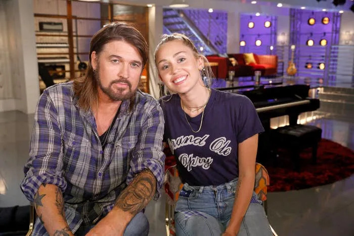 Billy Ray Cyrus and Miley Cyrus smiling, seated in a music-themed studio. Billy Ray wears a plaid shirt and has visible tattoos. Miley wears a Grand Ole Opry t-shirt and jeans