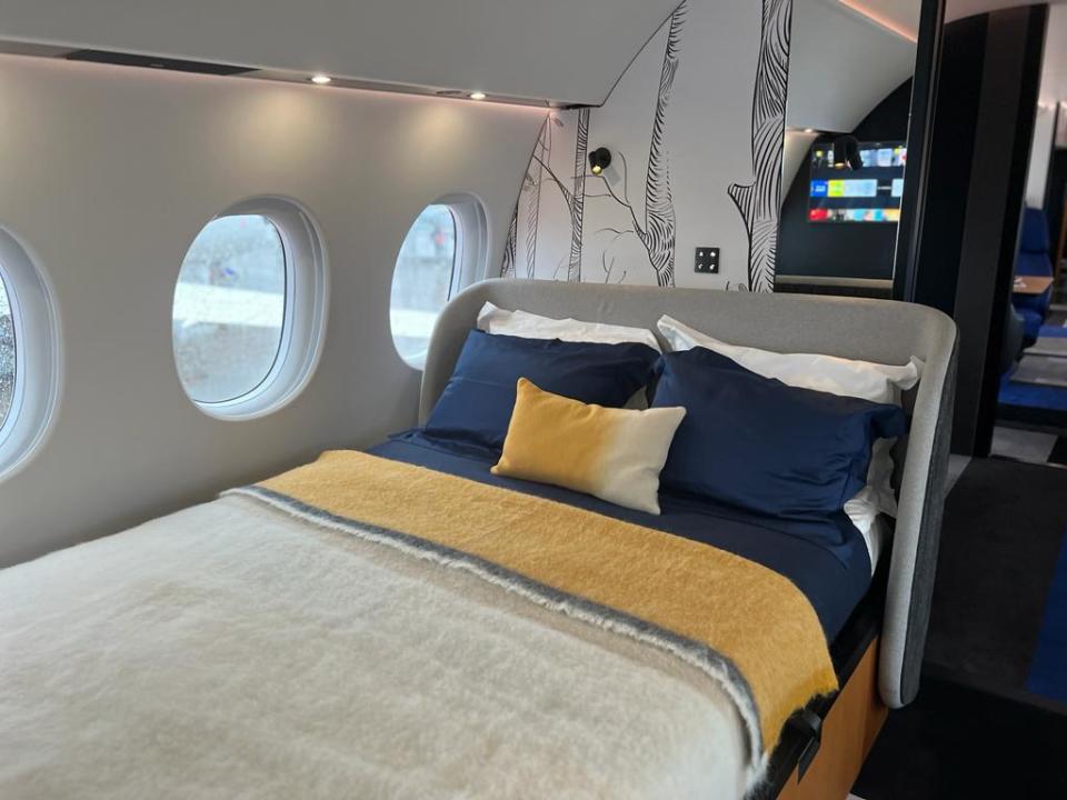 Closeup of the bed onboard a Falcon 10X, has a fluffy white comforter with yellow stripes and navy pillows