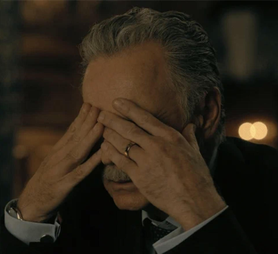 An older man with gray hair, mustache, and wearing a suit, appears stressed with his hands covering his face. He also wears a wedding ring