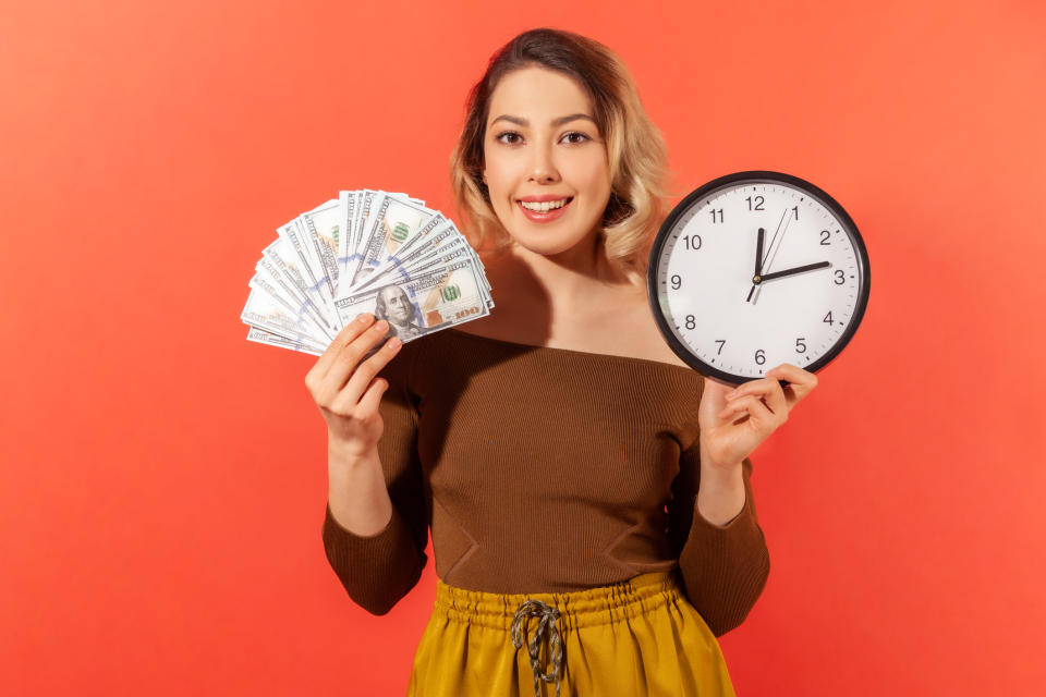 Happy woman holding dollar bills in one hand and a clock in the other. 