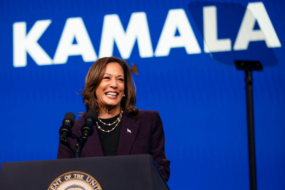 Kamala Harris delivering a speech at a podium, with "KAMALA" displayed in large letters behind her. She is smiling and wearing a suit jacket