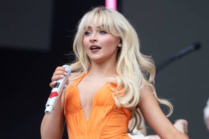 Sabrina Carpenter performing on stage, wearing a plunging neckline dress with voluminous hair, holding a microphone