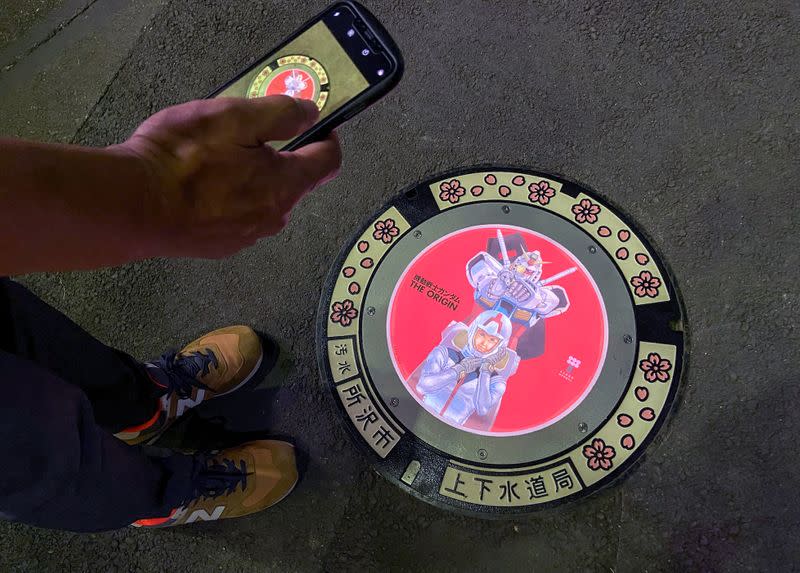 A passerby using a smartphone takes a photo of an illuminated manhole cover with designs of popular animation character Gundam, on the street in Tokorozawa, near Tokyo