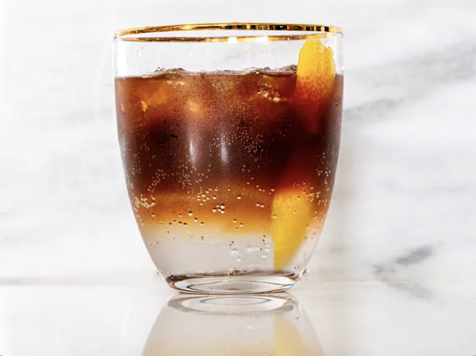 A glass filled with a dark, bubbly beverage, garnished with a lemon peel, is set against a marble background