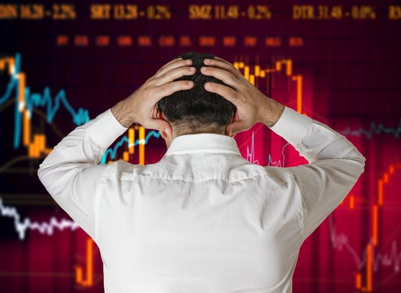 A man in a white button-down shirt facing a stock chart screen with hands on his head as if in frustration.