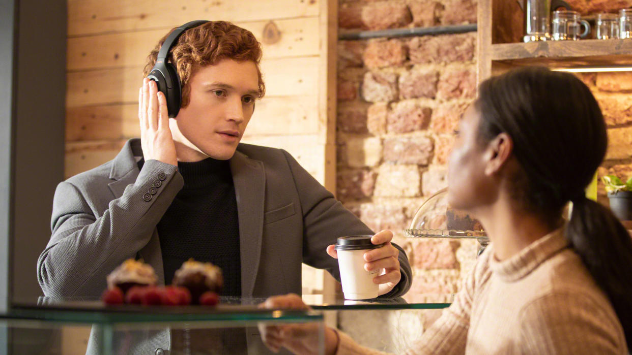  A man wearing headphones is holding a hand to his ear to activate Transparency Mode, while talking with café server. 
