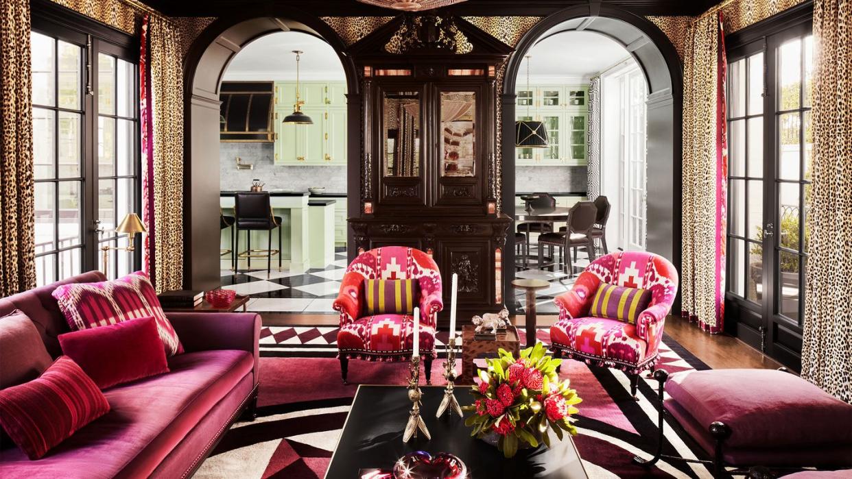 an antique buffet and two arched entryways frame a family room where a fuchsia velvet sofa, two fuchsia ikat armchairs, and two stools with rose colored cushions circle a cocktail table atop a patterned rug