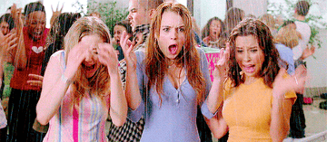 Three women express excitement while holding drinks, with a crowd cheering behind them