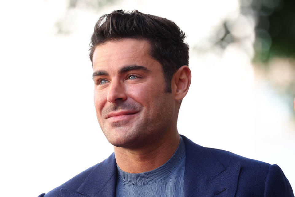 Zac Efron smiles at an event, wearing a suit jacket over a T-shirt