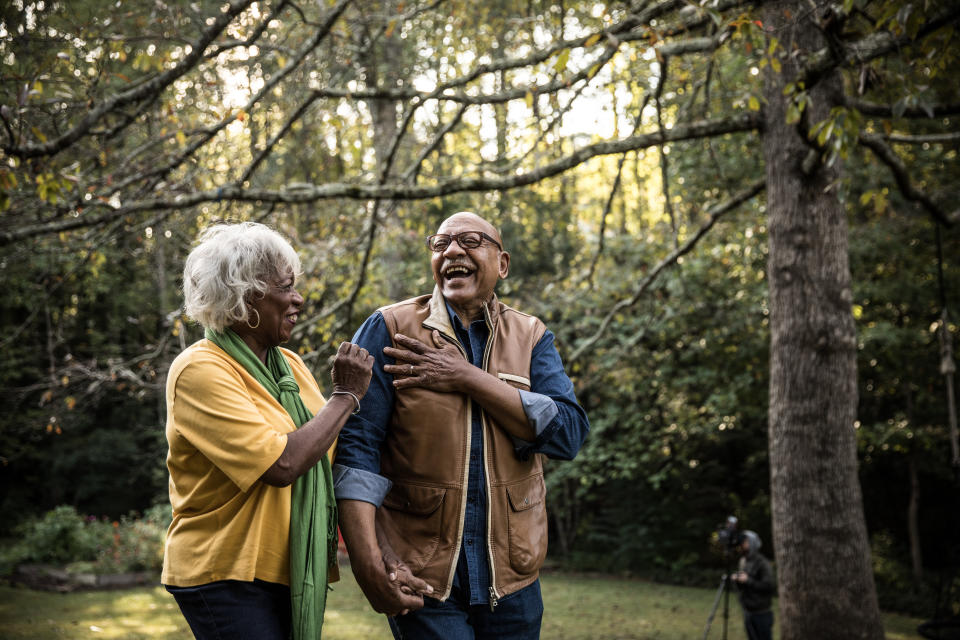 An elderly couple, both smiling and holding hands, stands in a wooded area with autumn foliage. They are dressed in casual clothing