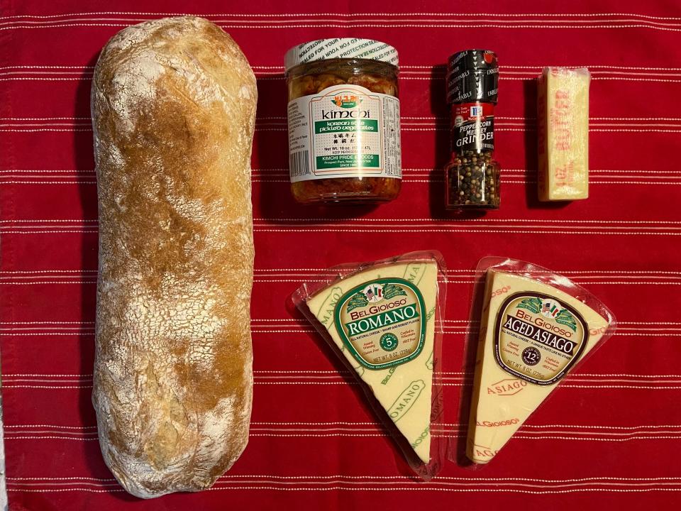 Ingredients laid out on a dishcloth including a ciabatta bread load, romano and asiago cheese, bottle of kimchi, peppercorns, and butter.