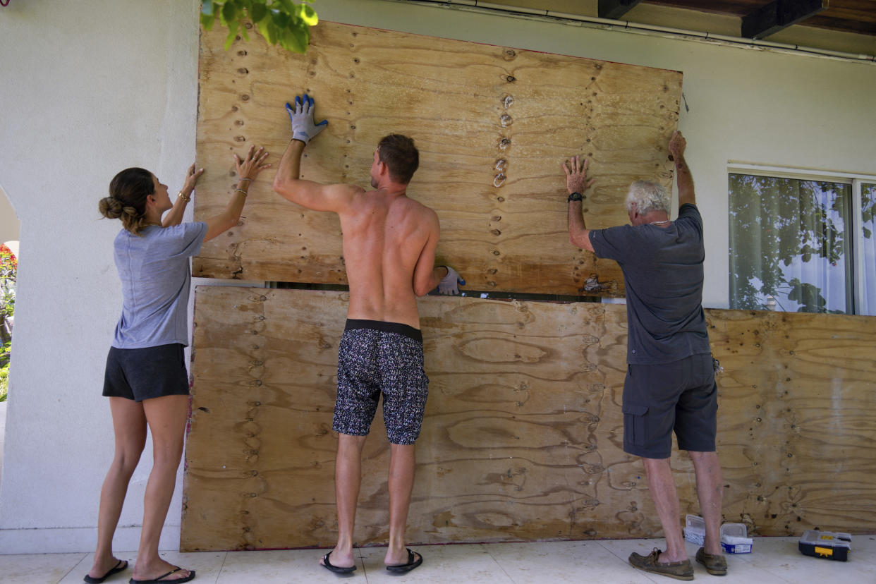 Three people hold plywood against the wall of a house.