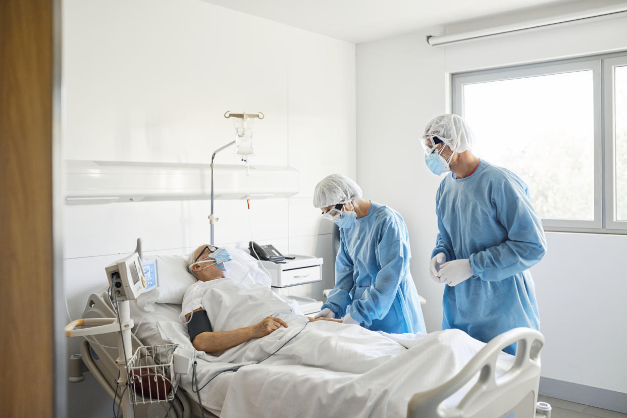 Male and female doctors in protective suits examining senior man lying on bed. Healthcare workers are examining patient in ward during COVID-19 outbreak. They are working in hospital during pandemic.