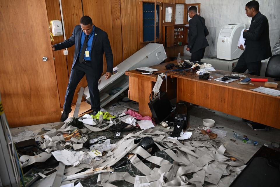 Planalto Presidential Palace security members gingerly step through a morass of papers and toppled furniture strewn over the floor in an office.