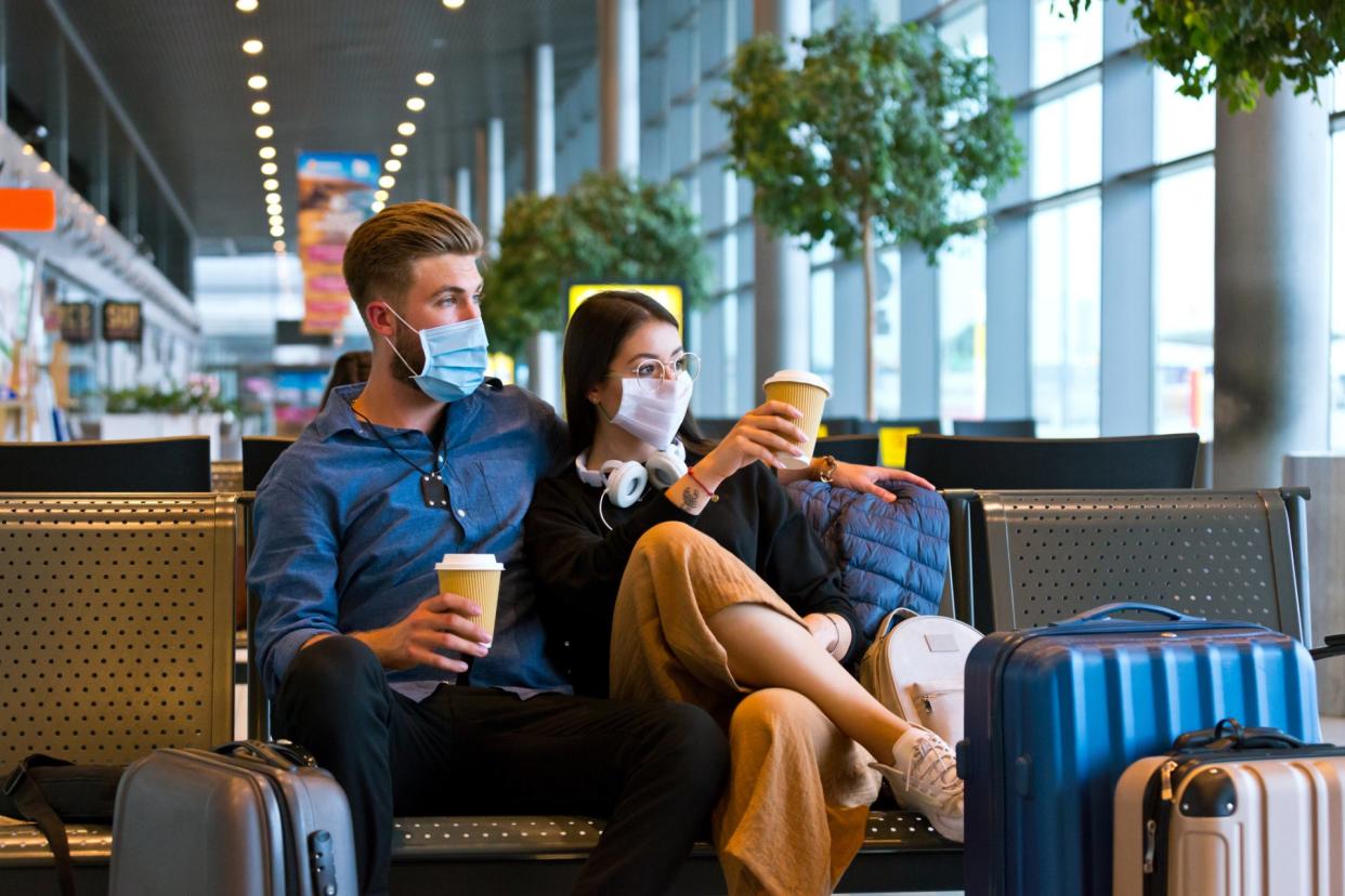 Young woman and man traveling by plane during COVID 19, wearing N95 face masks, sitting on bench with take away coffee in airport waiting area.