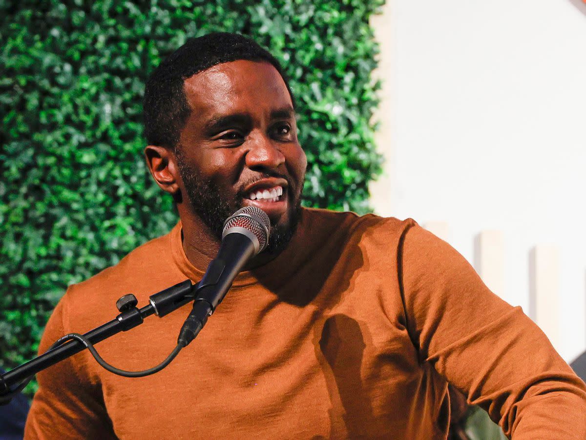 A Blakc man is smiling sitting at a microphone. He is wearing an orange shirt and smiling. A hedge can be seen behind him. 