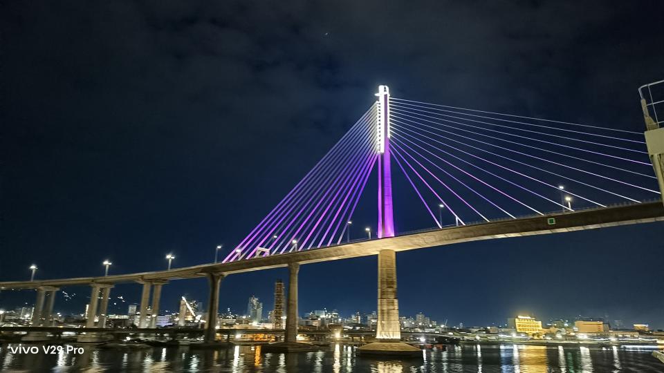 A night scene of a bridge with purple lights illuminating the water below