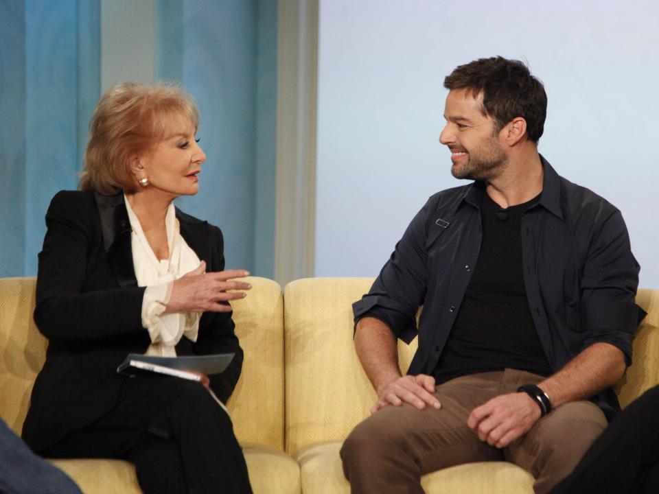 A woman with blonde hair in a black suit sits next to a man with black hair and a black jacket