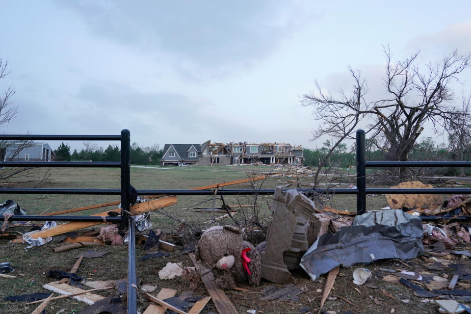 A view over to a large, wrecked residence shows a ripped-up iron fence and a huge plush bear sitting amid a pile of chewed-up metal and planks.