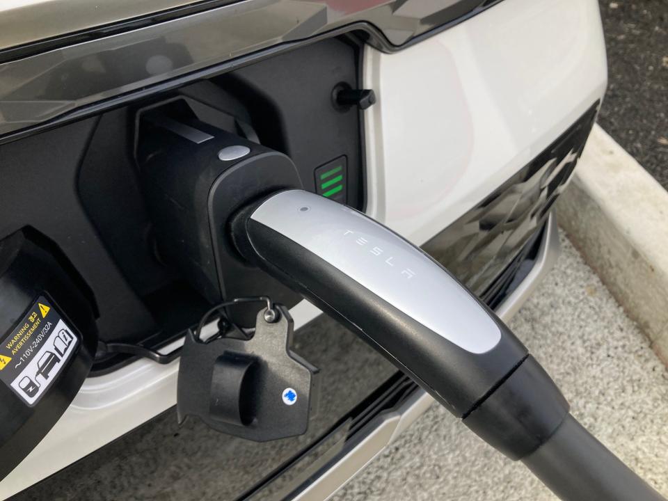 A close-up shot of a Tesla charging cable inserted into the charge port of a white Kia Niro EV electric SUV.