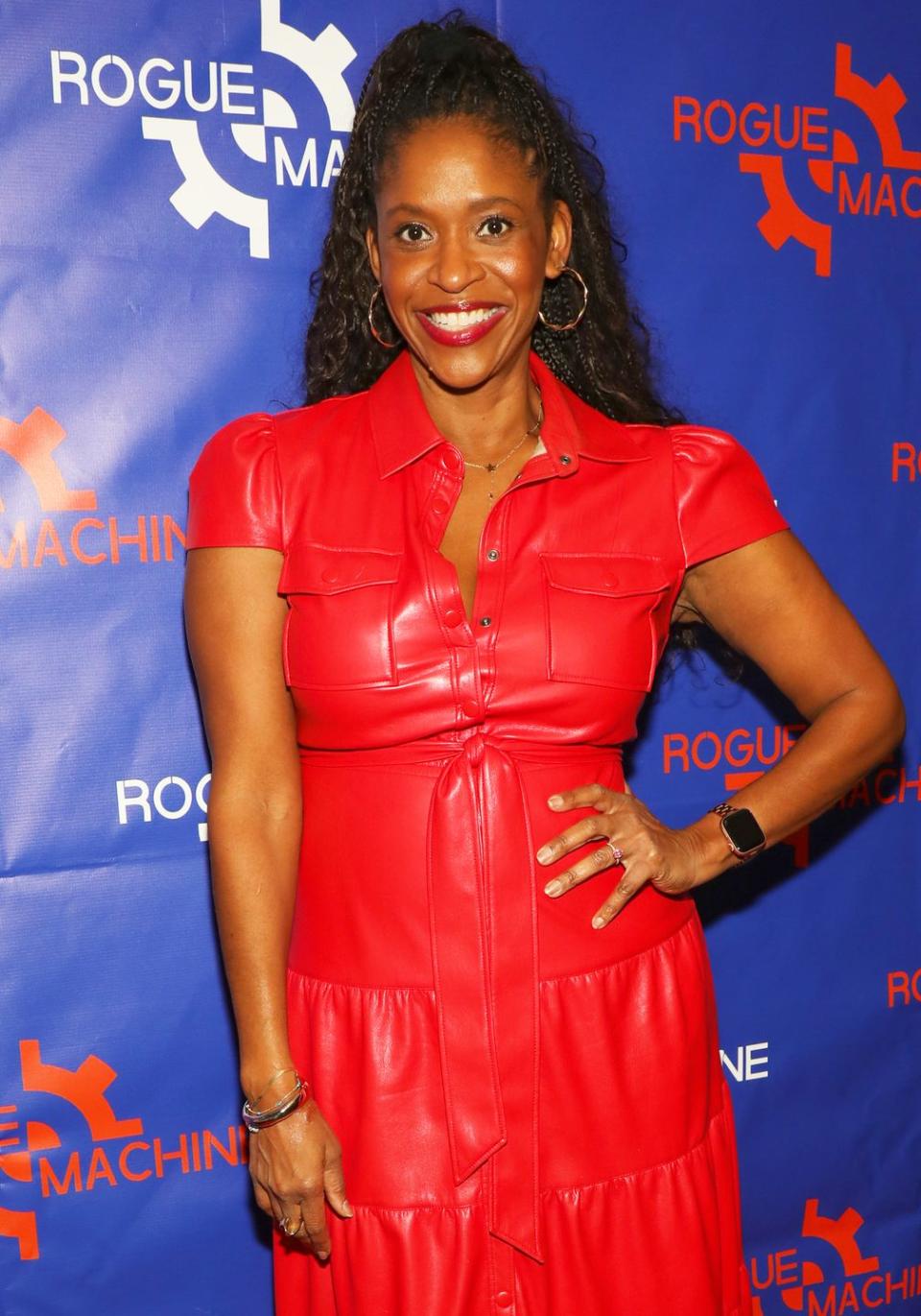 merrin dungey smiles for the camera in a red leather shirt dress and gold hopped earrings