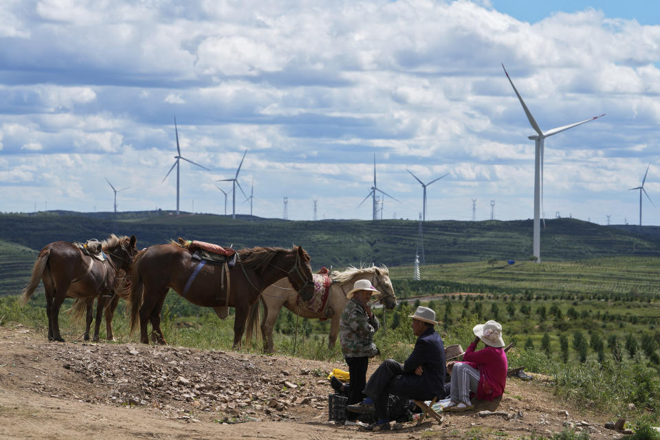 Villagers take a rest near horses on the grassland with wind turbines in the background in Zhangbei county, in north China's Hebei province on Aug. 15, 2022. The world's two biggest emitters of greenhouse gases are sparring on Twitter over climate policy, with China asking if the U.S. can deliver on the landmark climate legislation signed into law by President Joe Biden this week. (AP Photo/Andy Wong)