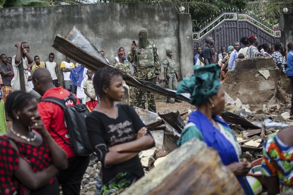 A soldier takes photos while women look at the remains of a house that was burnt down during communal violence ahead of elections in the Taouyah neighbourhood of Guinea's capital Conakry