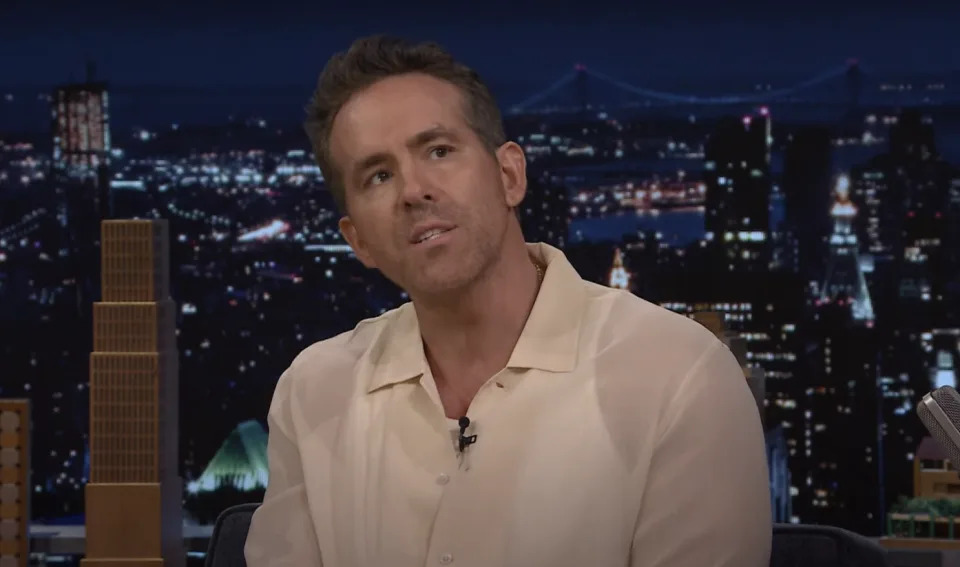 Ryan Reynolds is speaking on a talk show set in front of a cityscape backdrop at night. He is wearing a light, button-down shirt