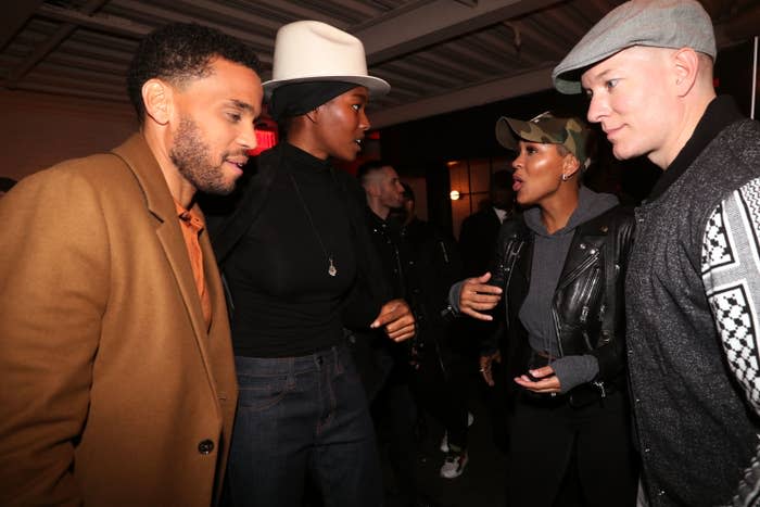 Four people, including Meagan Good (wearing a leather jacket and cap), having a lively conversation indoors at an event