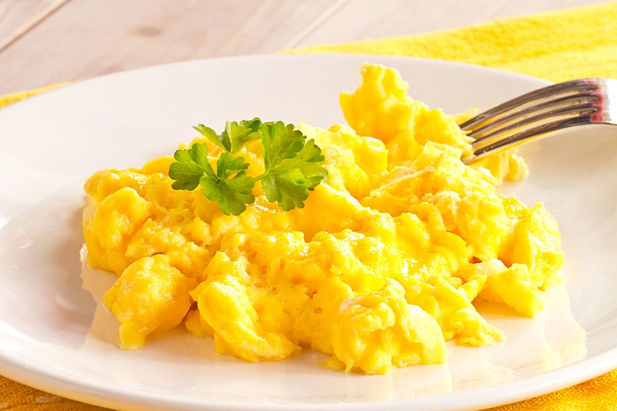 Focus on scrambled eggs on a white plate with a fork on a bright yellow napkin on a natural wooden table